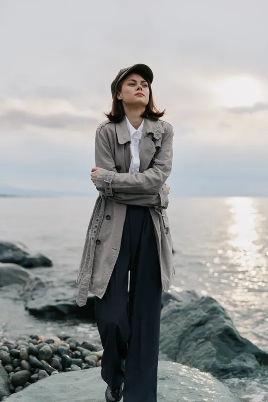 Young woman in a long patterned coat stands confidently on rocky shore at sunset. Calm sea and cloudy sky create a serene natural background for fashion concept.