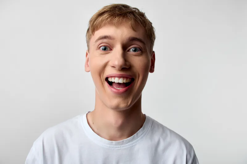 Excited, joyful, smiling man in a white shirt shows cheerful energy and expression in studio, capturing youthful happiness and candid closeup for visuals