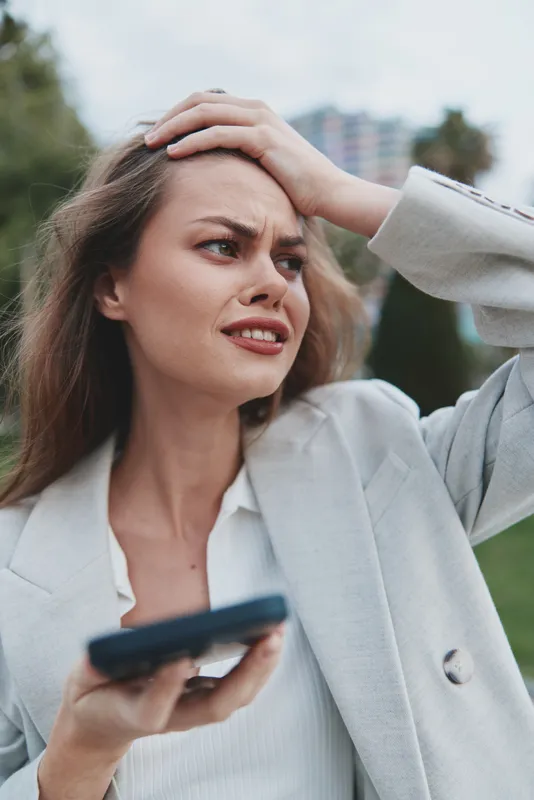 Young European woman stressed and worried holding smartphone outdoors in business casual jacket with upset facial expression, emotional tension, blurred city park background