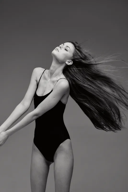 Black and white portrait of a young woman with long hair wearing a black swimsuit, posing gracefully against a plain background.