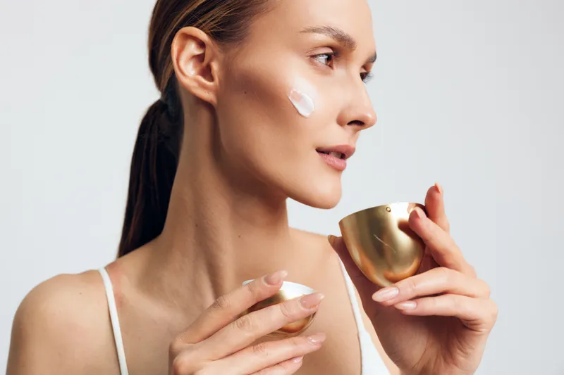 Cosmetic ritual unfolds as a woman applies face cream from a small jar in soft lighting, her profile serene and focused on the skincare moment.