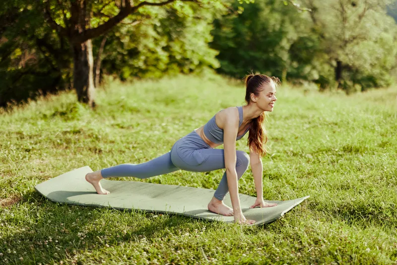 Young woman practicing yoga outdoors on a mat, showcasing flexibility and positivity amidst a serene green landscape, promoting wellness and mindfulness
