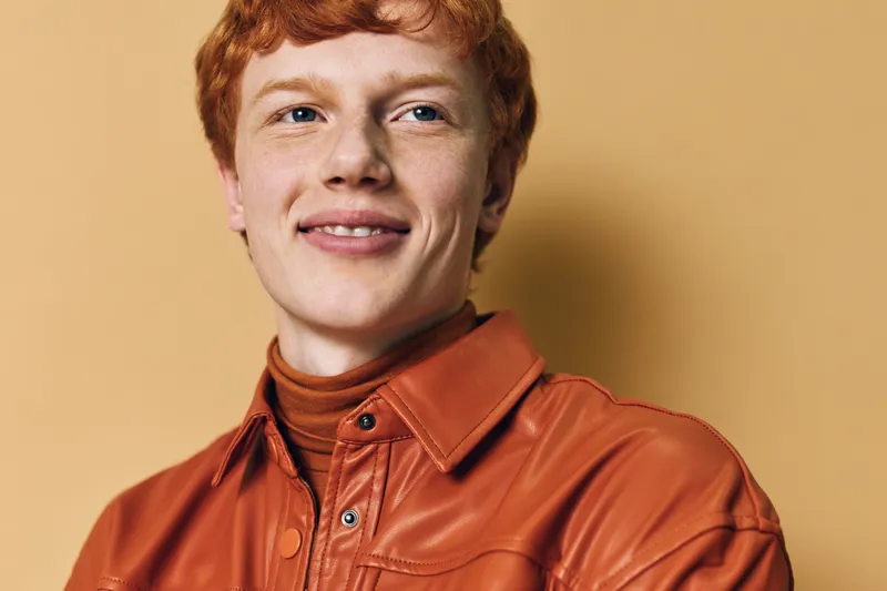 Young man with red hair wearing orange leather jacket and turtleneck smiling with slight smile on beige background. Male portrait and casual fashion style with confident look.