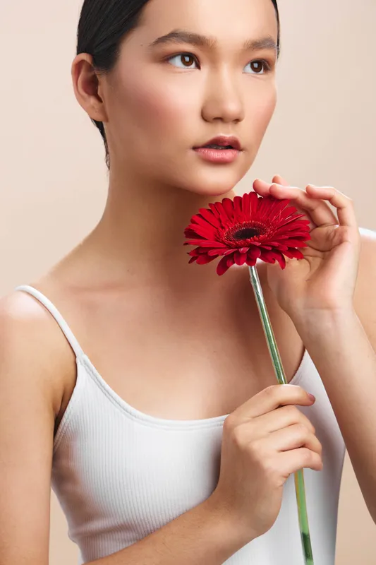 Asian woman holds a vibrant red flower near her face in a soft studio portrait. Calm expression, gentle light, and a serene mood highlight natural beauty.