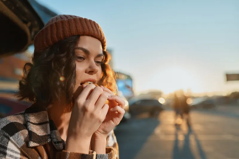 Woman portrait eating sandwich outdoors at golden hour, candid lifestyle capture with authenticity and mindful living mood, emotional storytelling in urban roadside scene.
