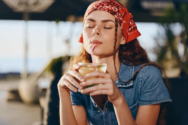 Lifestyle woman in modern boho-western denim street style enjoys a drink outdoors with relaxed expression and film color tones capturing casual summer vibe.