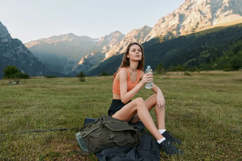 A cheerful young woman sits on a grassy field in a mountainous landscape, wearing an orange top and shorts, holding a water bottle, radiating a relaxed outdoor vibe.