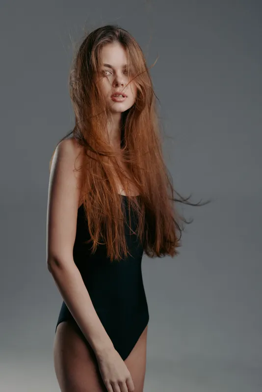 Young woman with long brown hair in black bodysuit posing against gray background