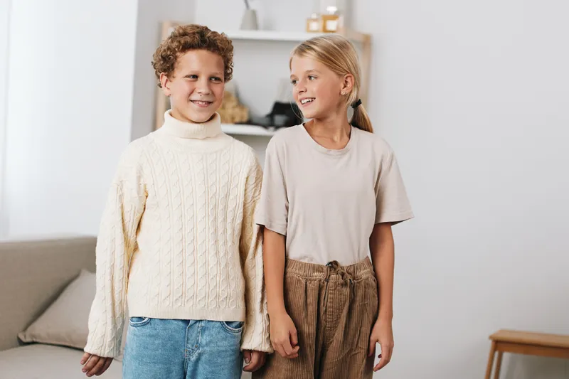 Two cheerful children stand indoors, smiling and close together, exuding warmth and friendship in a cozy living space with soft lighting and neutral decor.