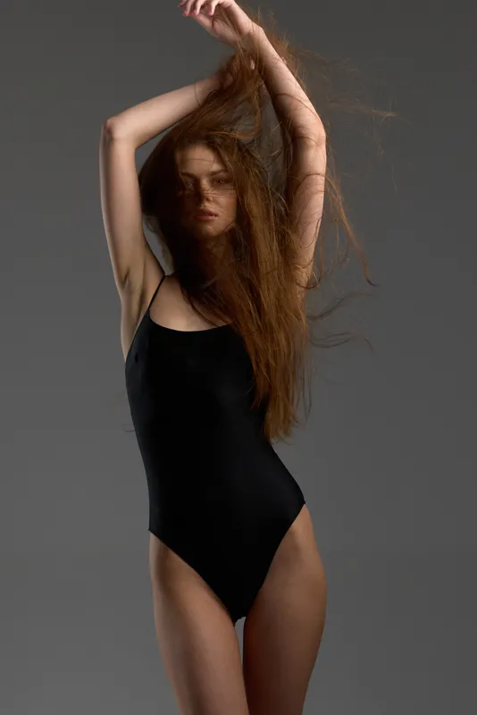 Woman with long hair in black swimsuit posing with arms raised against gray background