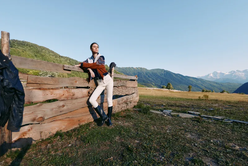 Woman leaning on wooden fence in mountain meadow wearing boots and casual outfit. Countryside nature scene with mountains, grass and wide outdoors landscape, relaxed mood.