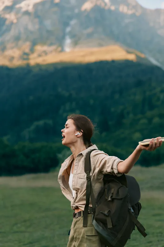 Cheerful woman outdoors, joyful expression, arms outstretched, backpack ready for adventure, natural landscape, open field, sunlight glow, casual outfit, carefree lifestyle