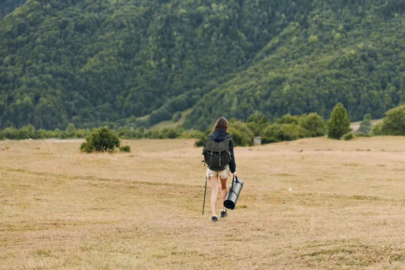 A lone hiker walks across a wide grassy field toward distant hills, carrying a backpack and trekking pole, exuding adventure and solitude in a serene outdoor landscape with quiet wind and expansive