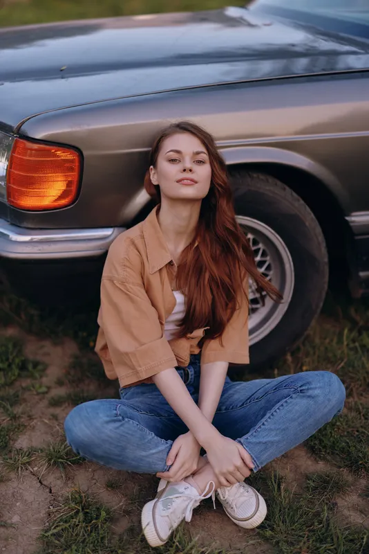 Young woman with long hair sitting beside a vintage car, dressed in casual outfit, expressing confidence and joy in a natural outdoor setting.