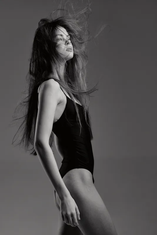 Black and white portrait of a tall woman with long hair in a black bodysuit, posing confidently with hair in motion against a plain background.