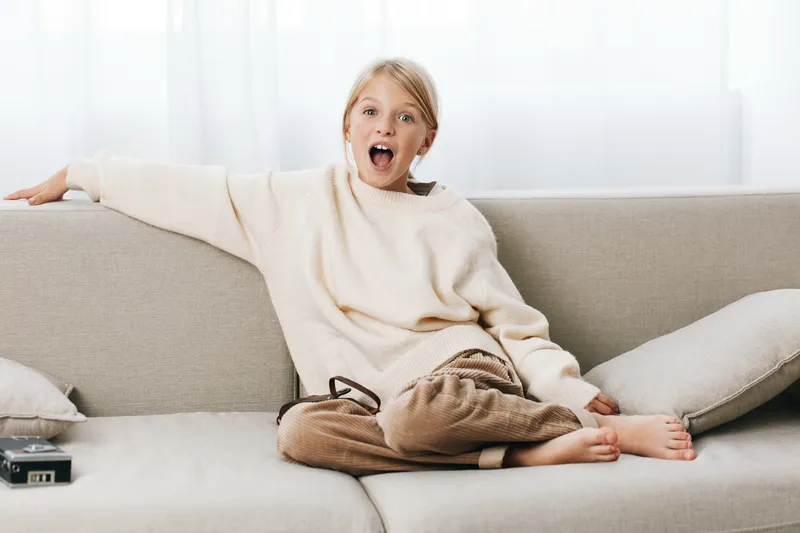 A surprised woman lounges on a light sofa in a cozy sweater, reacting with wide eyes and an open mouth, capturing a candid, intimate home moment indoors.