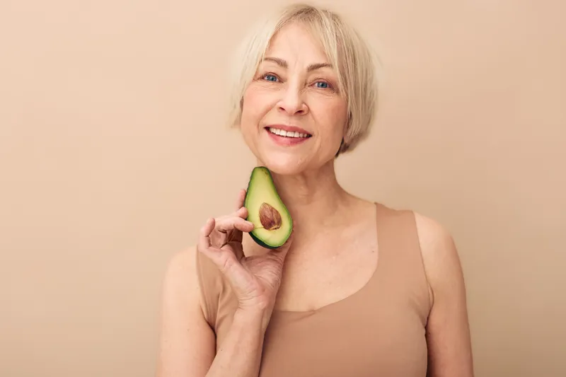 Smiling mature woman with short blonde hair holds half avocado near face on beige background. Concept of healthy lifestyle, nutrition, wellness, natural food, and positive aging.