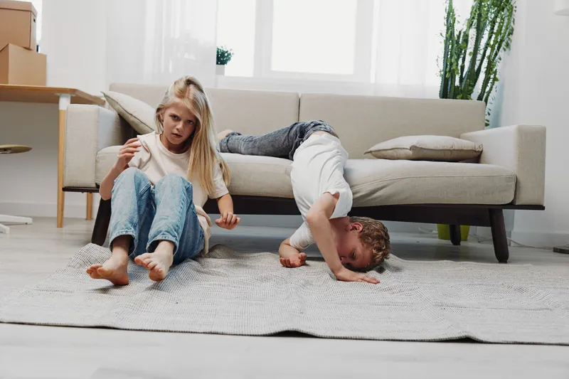 Two children play and relax in a bright living room with a modern sofa and natural light. The boy is lying on the sofa while the girl sits on the carpet. Concept of childhood and home.