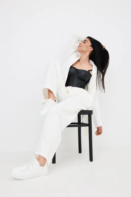 stylish woman in white pants and blazer poses seated on black chair with casual sneakers against plain white background, showcasing modern fashion and relaxed elegance