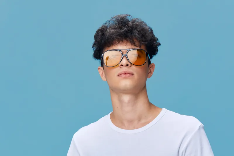young man wearing reflective sunglasses and white t-shirt stands confidently against blue background, showcasing stylish casual summer look and modern hairstyle.