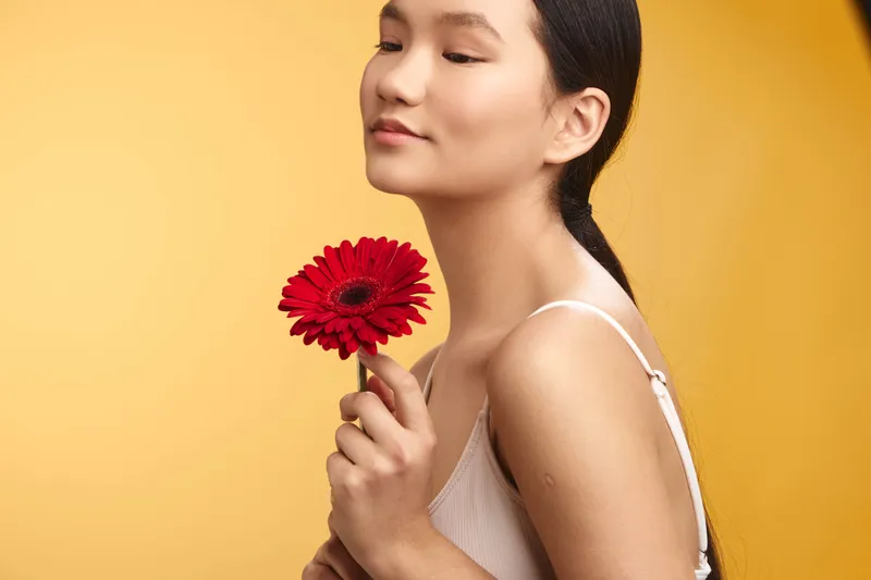 asian woman stands before a warm orange backdrop, holding a red flower with a gentle smile. soft lighting highlights graceful features, creating a serene, elegant mood.