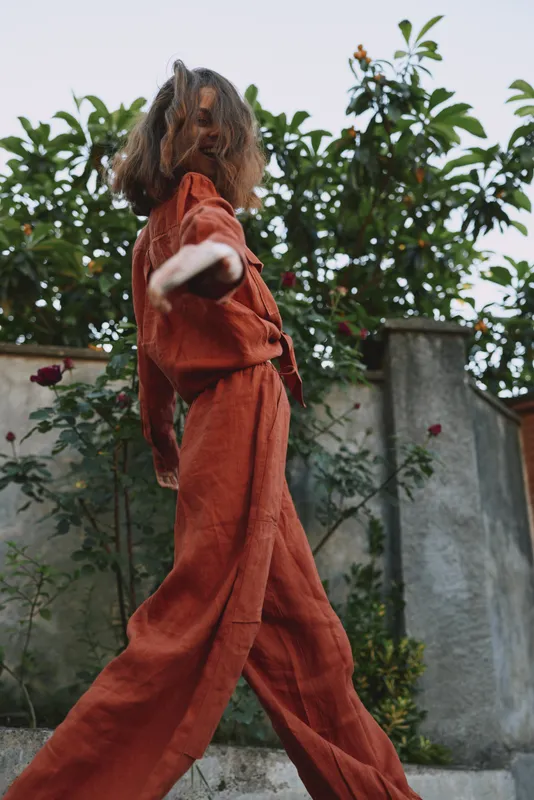 Young woman in an orange jumpsuit strolls outdoors with hair loose, sunlit garden behind her, conveying a confident carefree lifestyle moment