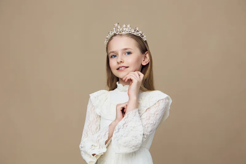 Young girl with a crown wearing a white dress, showcasing a thoughtful expression against a neutral beige background, perfect for themes of childhood and celebration