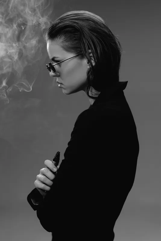 Black and white portrait of a thoughtful woman smoking with sunglasses in stylish outfit and slicked back hair.