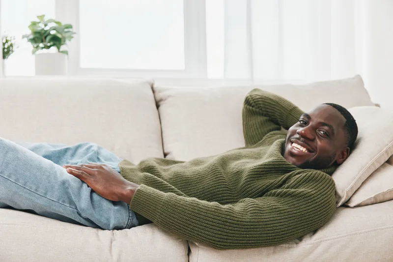 Smiling young African man relaxing on a couch in a cozy living room, wearing a green sweater, capturing a warm and inviting atmosphere