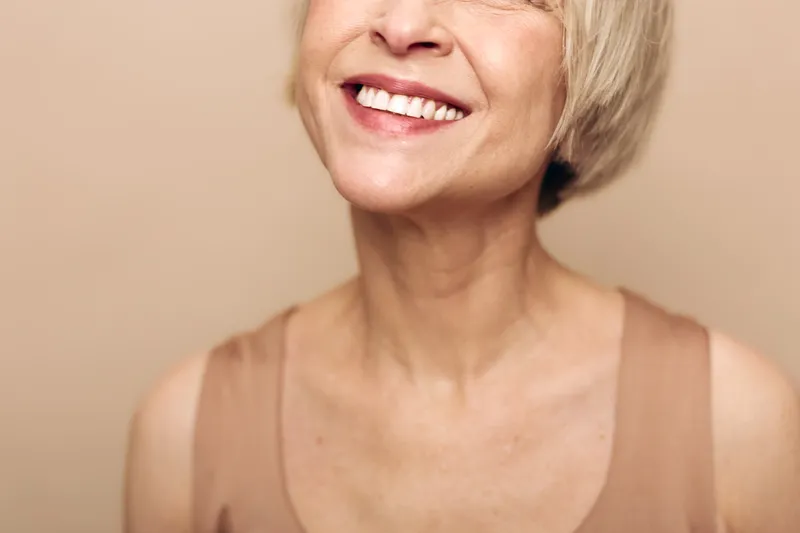 Close up of smiling mature woman with short blonde hair and smooth skin wearing beige top against neutral background. Concept of happiness, confidence, beauty, aging, and natural look.