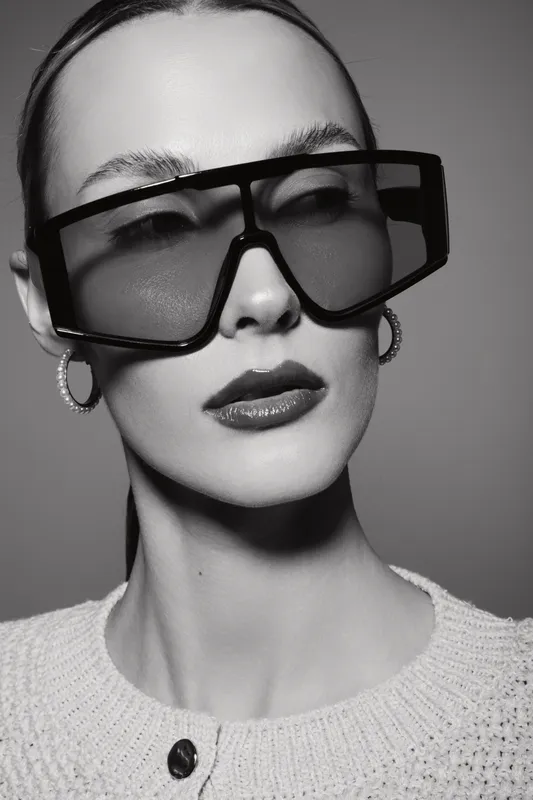 Stylish white woman with pale skin wearing large futuristic sunglasses and hoop earrings poses in black and white studio portrait.