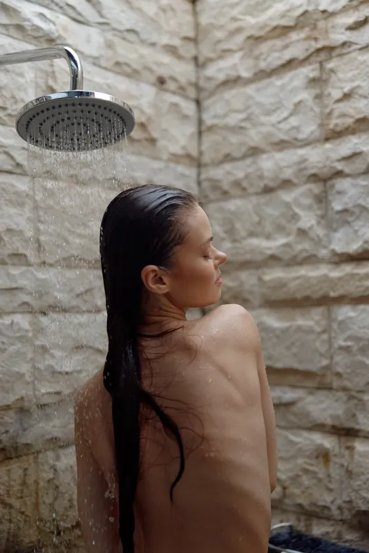 naked woman under outdoor shower with wet long hair on natural stone wall background, serene mood, natural light, relaxing moment, summer setting