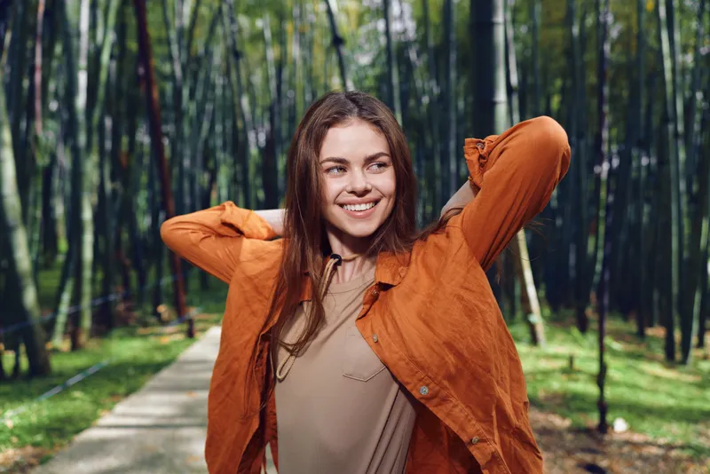 Woman smile forest bamboo path jacket portrait outdoors happy in orange jacket on a sunlit walkway, candid portrait of a young woman enjoying nature, relaxed travel and casual style.
