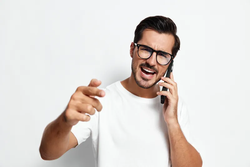 Expressive young man wearing glasses talking on mobile phone, gesturing with hand, showcasing excitement and engagement, isolated on plain solid white background.