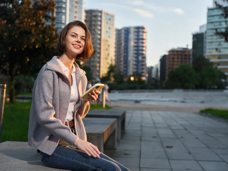 A cheerful woman sits outdoors in an urban park, smiling while texting on her phone, enjoying a calm moment as the city skyline frames a bright, hopeful scene.