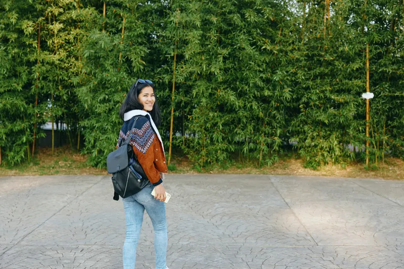 Woman with backpack walking on a paved street beside green trees, casual jacket and jeans, smiling and turning toward the camera, outdoor lifestyle moment