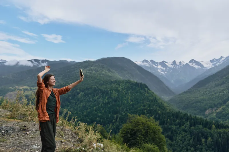 A traveler stands on a mountain overlook, taking a selfie as valley and snow capped peaks stretch into the blue sky, capturing outdoor adventure and expansive scenery.