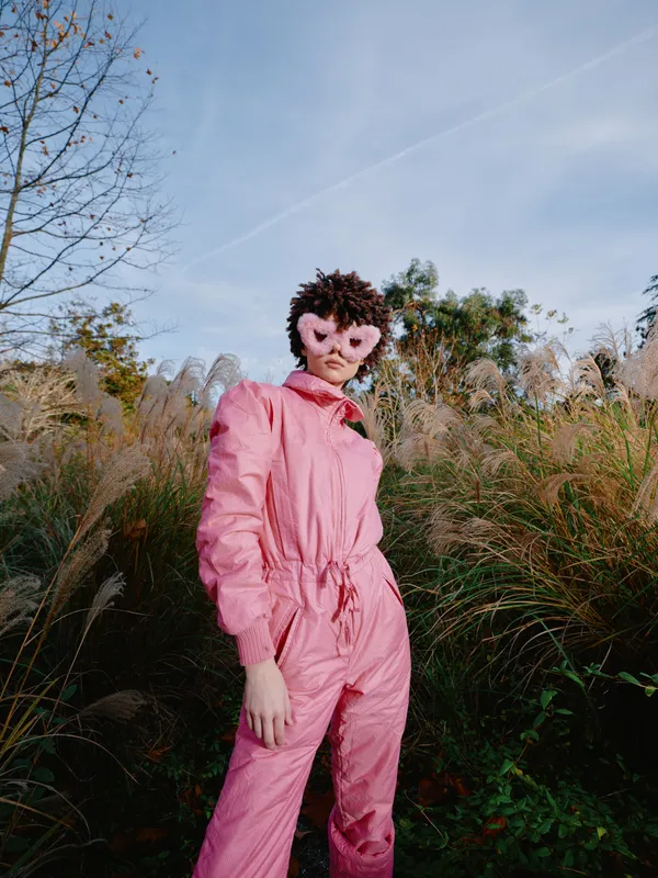 Pink jumpsuit wearing person with animal mask standing in a grassy field under a clear blue sky, creating a surreal fashion portrait outdoors for photography