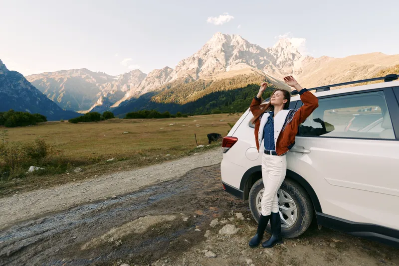 Woman leans against white car beside gravel road with mountains, landscape and nature view during roadtrip. Travel outdoors scene with relaxed pose and scenic adventure.