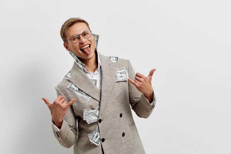 young caucasian man celebrating success wearing light brown coat with scattered dollar bills showing rock sign excited isolated on white background symbolizing freelance jobs and cryptocurrency