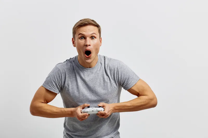 Excited man playing video game holding gamepad in hands wide-eyed surprised facial expression casual grey t-shirt indoor studio shot. People entertainment leisure lifestyle concept