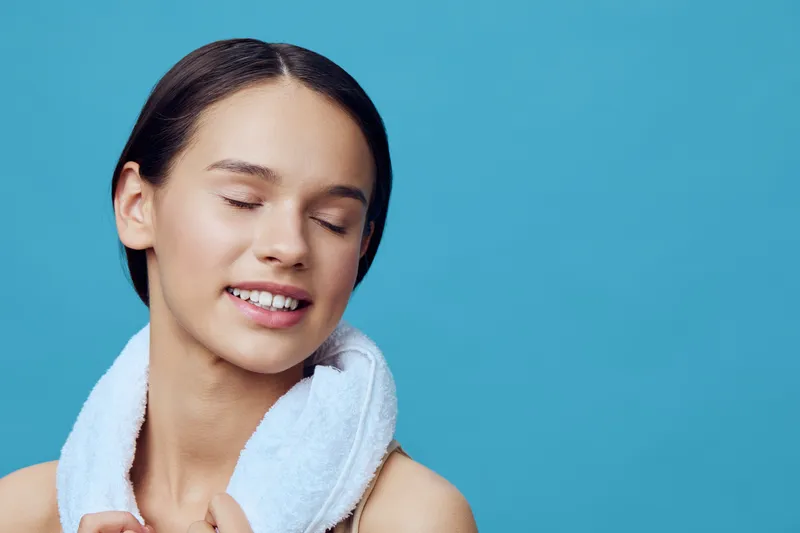 Relaxed young woman with a towel on her shoulders, smiling with closed eyes, showcasing beauty and wellness against a soft blue background