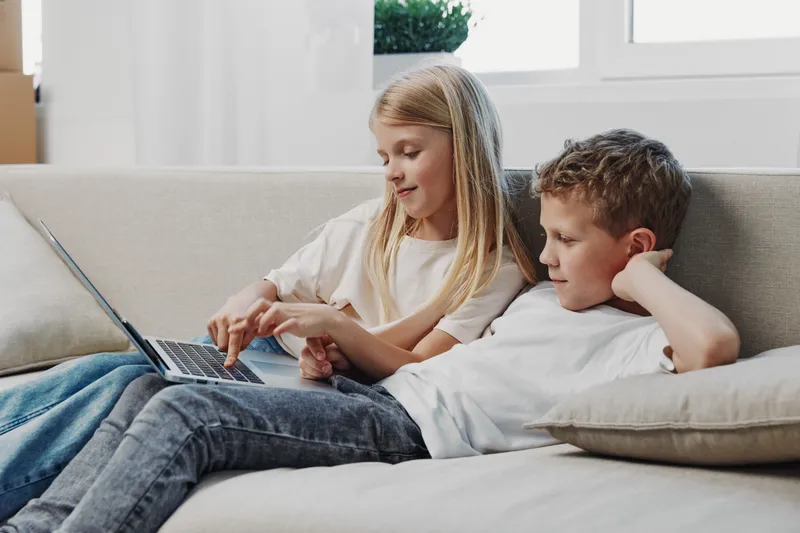Two children relax on a couch using a laptop together in a bright room, focused on the screen. Concept of family bonding, technology, and childhood learning.