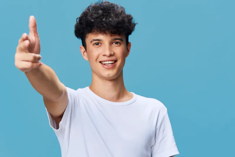 smiling man in white t-shirt reaching out hand with curly hair standing against blue background, cheerful young adult extending greeting with friendly expression