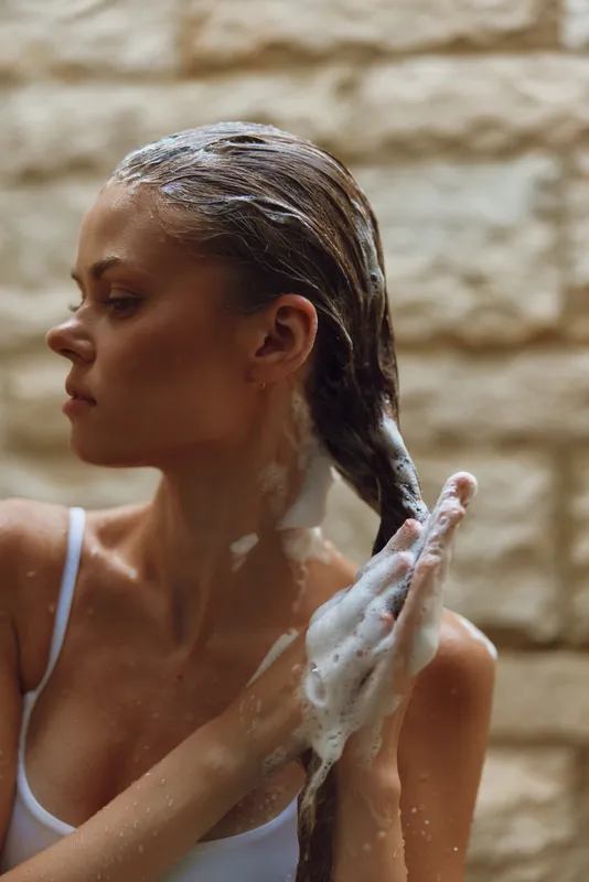Woman washing long hair with shampoo foam, showing wet braided hairstyle and relaxed mood indoors against stone wall background.