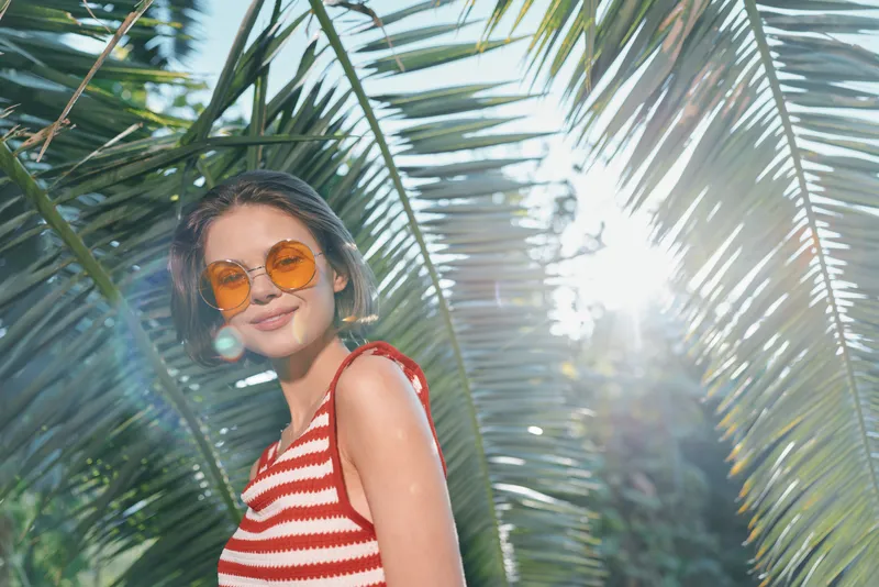 A cheerful woman wearing a striped red and white top and orange sunglasses stands among palm leaves in a sunny tropical setting, radiating relaxed confidence and summer joy.