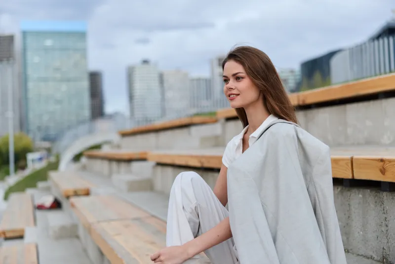 Woman in light gray jacket sitting on urban steps with modern cityscape background smiling confidently in casual fashion style