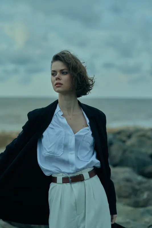 Fashion model on a windy coast wearing white shirt and white pants under a black blazer, belt, and flowing hair against a moody sea backdrop