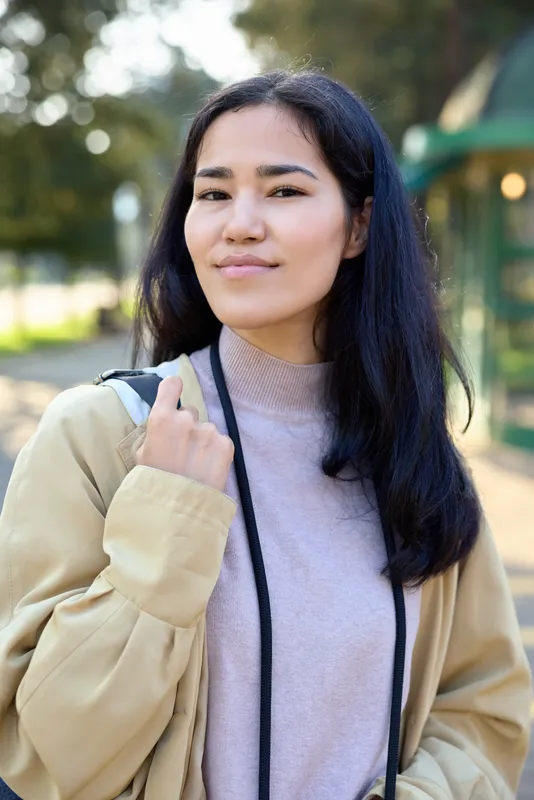 A woman wearing a beige cardigan and soft sweater stands outdoors in a sunny park, carrying a shoulder bag, smiling and relaxed for a casual lifestyle shot suitable for fashion and everyday imagery