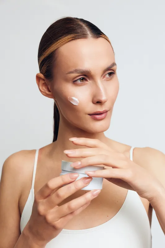 Young woman with clear skin applies moisturizing cream on her face holding a jar of skincare product isolated on white background with natural light and minimal makeup.